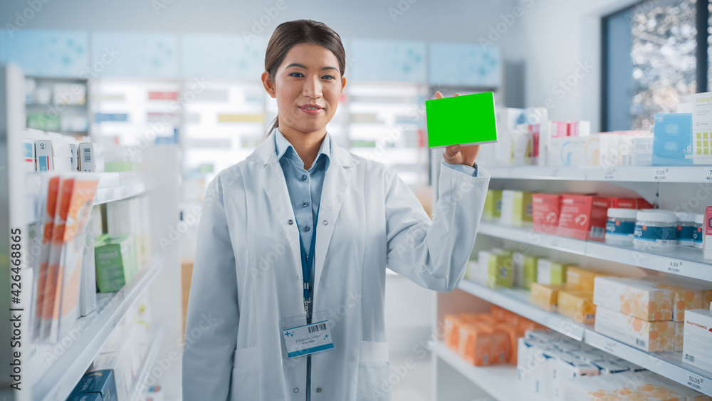 Pharmacy Drugstore: Portrait of Professional Asian Female Pharmacist Holding Mock-up Template Medicine Package with Green Template, Looking at Camera, Smiling. Specialist Recommending Best Product
