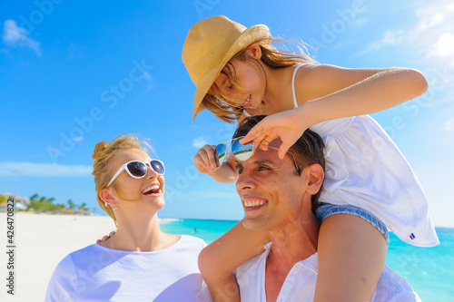 Family at the beach, dressed in white outfits, daughter sitting on father's shoulder and holding his sunglasses while mother looks on
