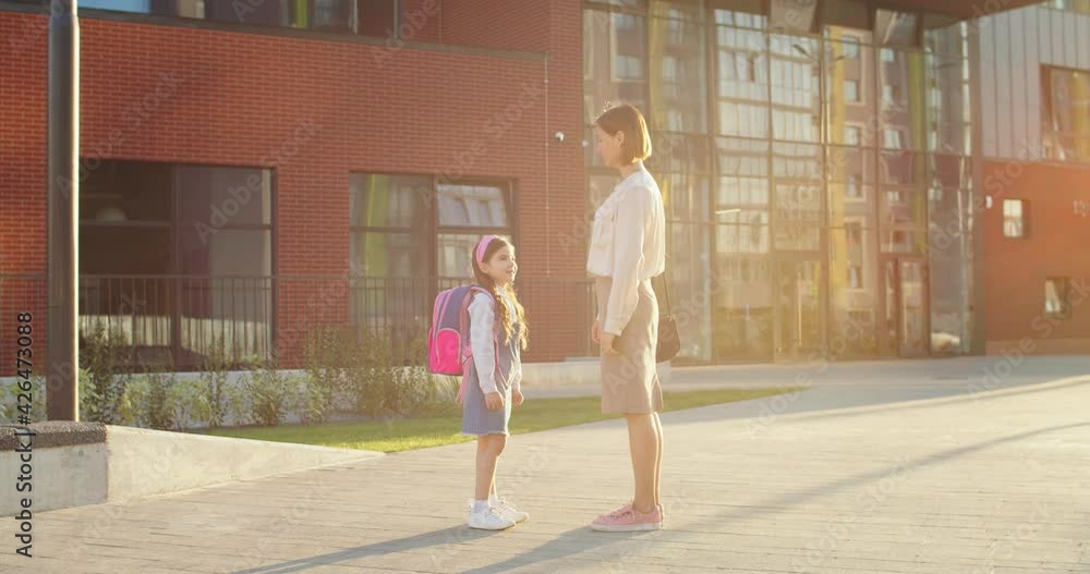 Caucasian happy beautiful woman talking to cute small daughter near school on sunny day. Little joyful girl chatting with mother in good mood while standing outdoors. Back to school concept Caucasian happy beautiful woman talking to cute small daughter near school on sunny day. Little joyful girl chatting with mother in good mood while standing outdoors. Back to school concept
