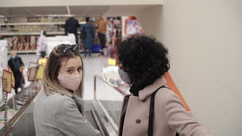 Young women with a protective masks taking the escalator stairs in a shop in the mall, looking in the camera. Shopping during covid pandemic Young women with a protective masks taking the escalator stairs in a shop in the mall, looking in the camera. Shopping during covid pandemic