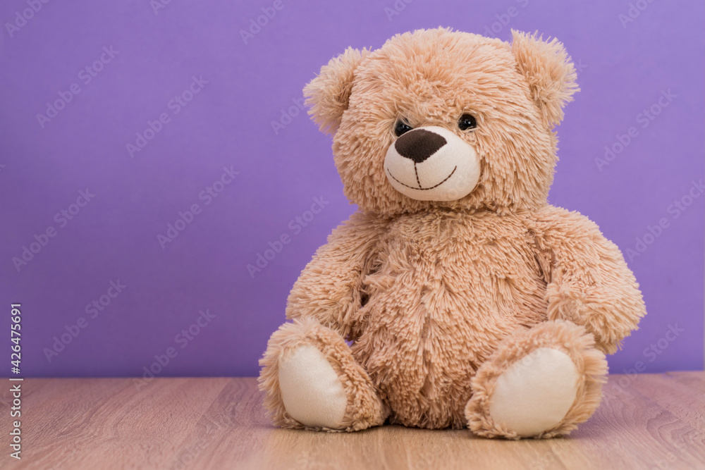 Fototapeta premium Smiling teddy bear sits on the table against the background of a purple wall. Space for text, copy space