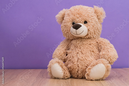 Smiling teddy bear sits on the table against the background of a purple wall. Space for text, copy space