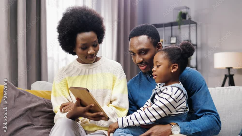 Portrait of cute African American family parents and little girl kid sitting on couch in living room at home and tapping on tablet teaching daughter using educative app on device, parenting concept Portrait of cute African American family parents and little girl kid sitting on couch in living room at home and tapping on tablet teaching daughter using educative app on device, parenting concept