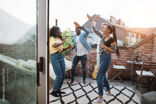 Multi ethnic group of friends dancing and relaxing on rooftop. Group young hipsters drinking alcohol and chatting. Carefree weekends.