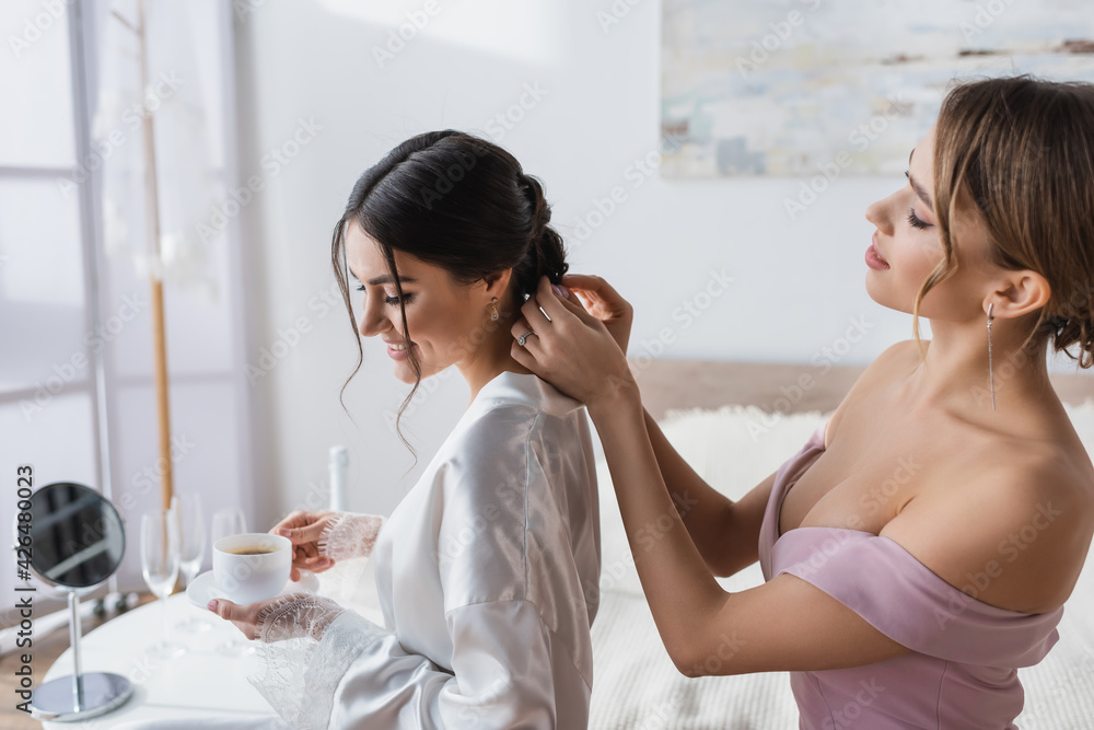 young woman making hairstyle to smiling bride holding coffee in bedroom.