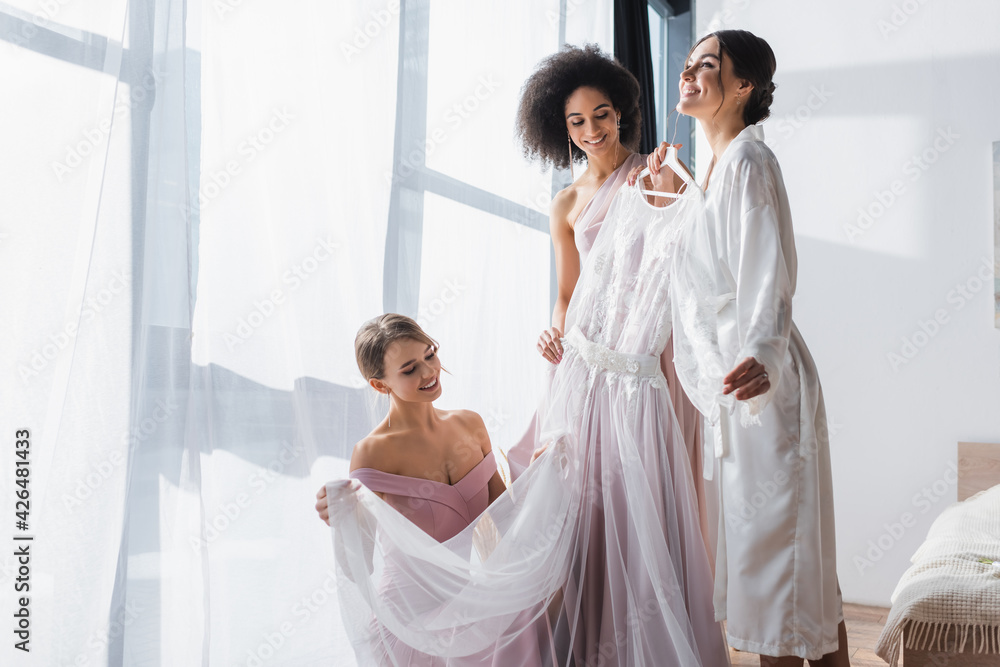 Obraz premium happy woman holding wedding dress near interracial bridesmaids.