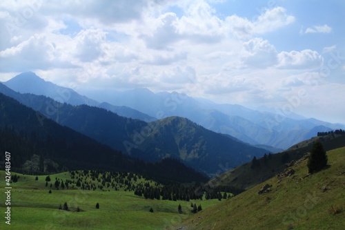 The high-mountainous green valley of Kok-Zhailau with ridges, a grassy glade, a forest is growing, a series of ridges goes into the distance, the sky is with clouds, summer, sunny