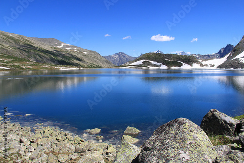 Mirror of a high-mountain lake in Altai, surrounded by mountain ridges and peaks with snow, stones on the shore of the lake, sky with clouds, summer, sunny