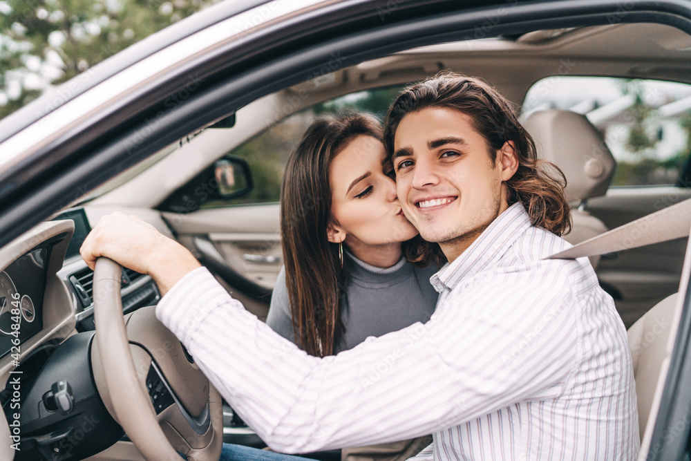 Happy smiling man behind the wheel of his car, fastened his seat belt, he smiles at the camera, next to him a young girl kisses him on the cheek