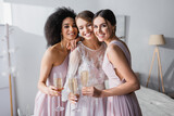 cheerful interracial bridesmaids holding champagne glasses near happy bride in bedroom.