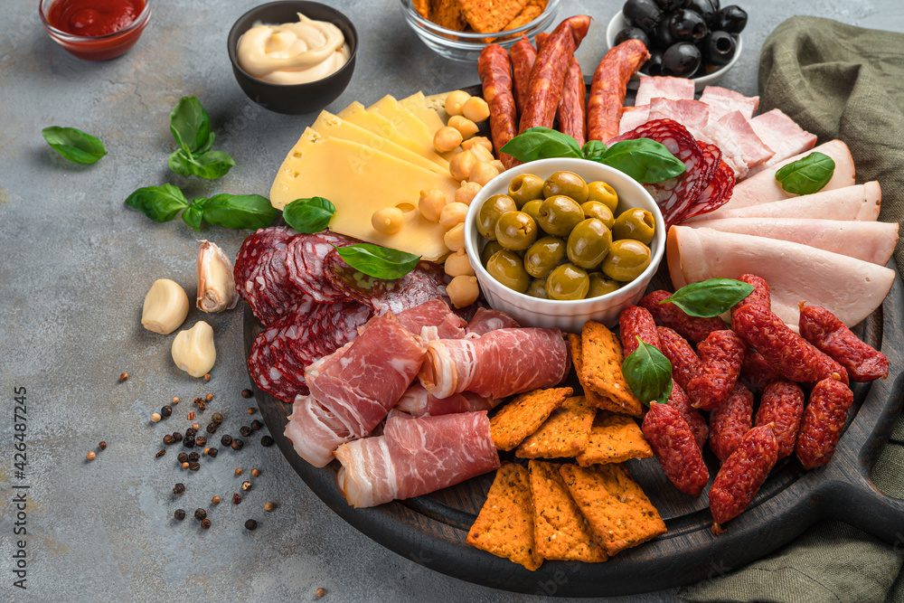Round board with sausage and cheese slices, olives, basil and sauces on a dark gray background. Round board with sausage and cheese slices, olives, basil and sauces on a dark gray background.
