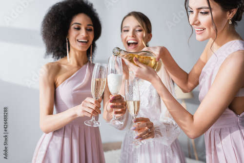 young woman pouring champagne near african american friend and bride on blurred background.