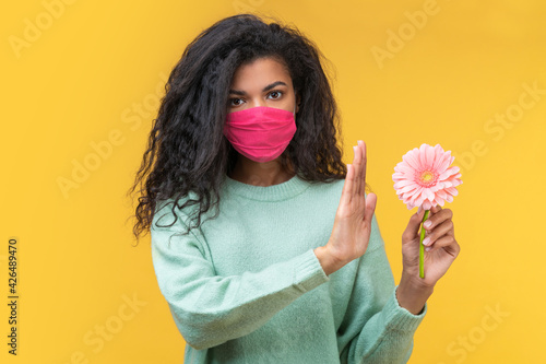 Studio portrait of african american girl in protective face mask unwilling to take herbera daisy flower because of pollen allergy photo