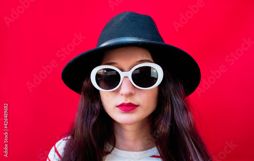 Portrait of a young girl with sunglasses and hat on red background © Nedrofly