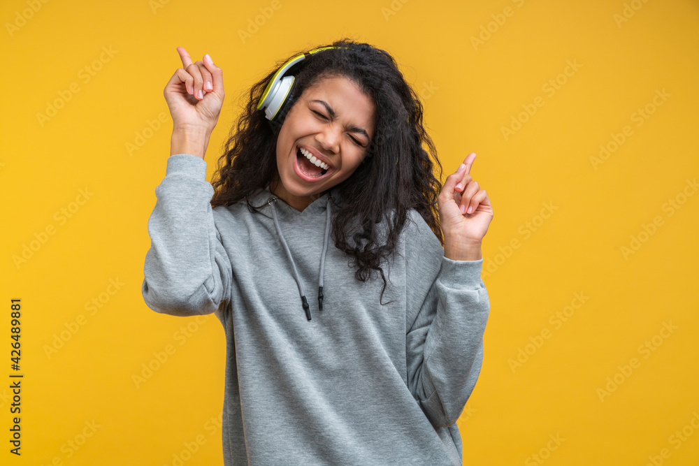 Portrait of dark skinned hipster girl wearing grey basic hoodie and wireles headphones enjoying listnening to music and dancing isolated over bright colored orange yellow background Portrait of dark skinned hipster girl wearing grey basic hoodie and wireles headphones enjoying listnening to music and dancing isolated over bright colored orange yellow background