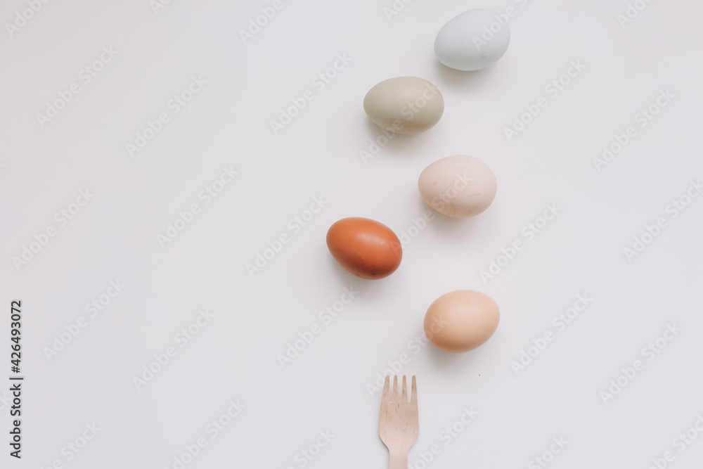 Chicken eggs of natural shades and colors and wooden fork, bamboo cutlery on white background. Healthy organic food, natural farmer's products, eco-friendly 
