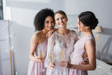 joyful bride looking at camera while holding champagne together with interracial friends.