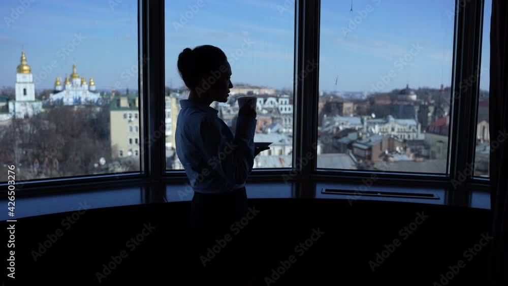 Silhouette of businesswoman drinking tea and admiring scenic view on city historic buildings outside office window. Mixed race woman chief relaxing with cup of hot drink by panoramic window Silhouette of businesswoman drinking tea and admiring scenic view on city historic buildings outside office window. Mixed race woman chief relaxing with cup of hot drink by panoramic window