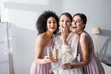 laughing bride covering mouth with hand near interracial bridesmaids with champagne glasses.