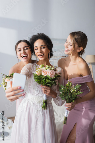 african american bride taking selfie with cheerful bridesmaids at home.