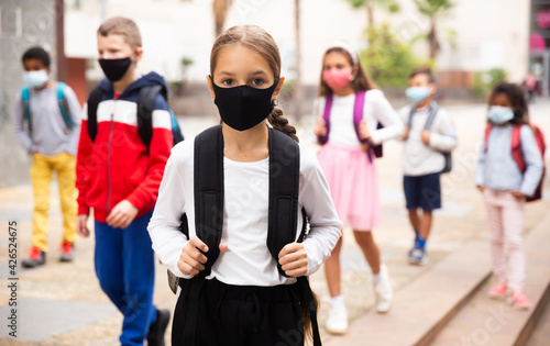 Portrait of positive schoolgirl in mask standing near school, children on background