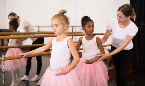 Two little girls practicing choreographic elements on ballet barre with help of teacher in dance school