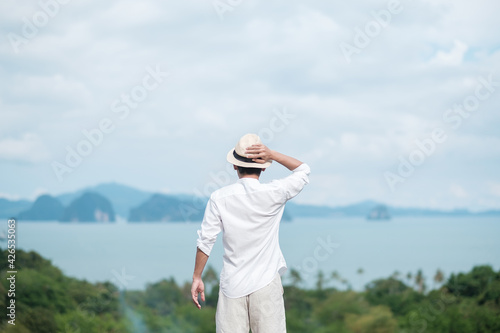 Happy traveler man in white shirt enjoy Beautiful view, alone Tourist with hat standing and relaxing over ocean. travel, summer and vacation concept