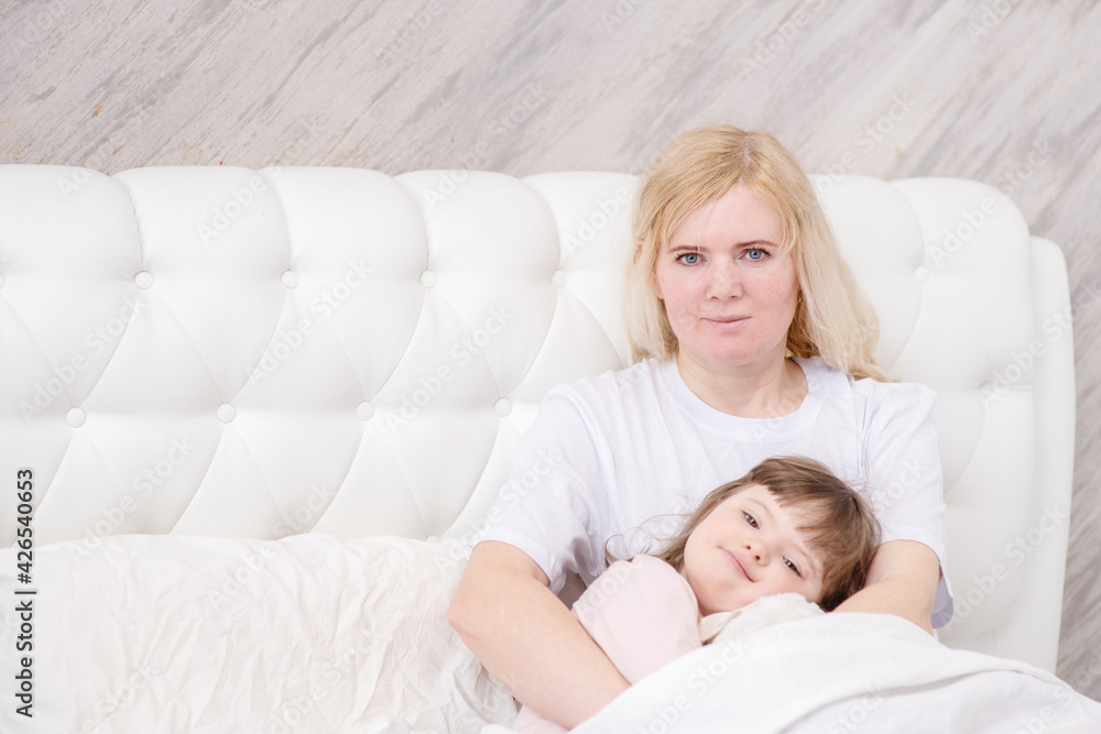 Mom hugging a girl with down syndrome holding a teddy bear at home in the bedroom on the bed. Ordinary childcare in a family for children with disabilities Mom hugging a girl with down syndrome holding a teddy bear at home in the bedroom on the bed. Ordinary childcare in a family for children with disabilities