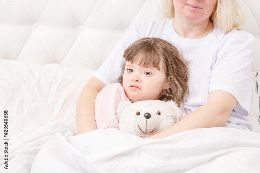Mom hugging a sleeping girl with down syndrome holding a teddy bear at home in the bedroom on the bed. Ordinary childcare in a family for children with disabilities Mom hugging a sleeping girl with down syndrome holding a teddy bear at home in the bedroom on the bed. Ordinary childcare in a family for children with disabilities