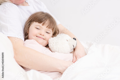 Mom hugging a sleeping girl with down syndrome holding a teddy bear at home in the bedroom on the bed. Ordinary childcare in a family for children with disabilities