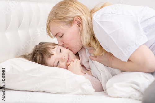 Mom kissing a girl with down syndrome in the bedroom on the bed while going to bed. Ordinary childcare in a family for children with disabilities