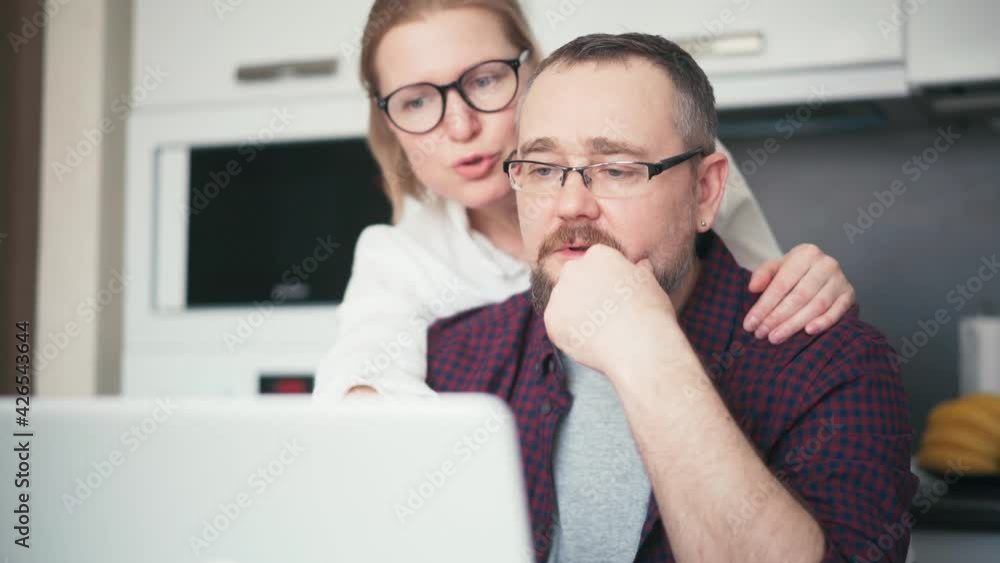 A serious couple husband and wife talking while sitting in the kitchen and looking at a laptop screen. Married spouse checking banking online application on a laptop, receiving bad news notification. A serious couple husband and wife talking while sitting in the kitchen and looking at a laptop screen. Married spouse checking banking online application on a laptop, receiving bad news notification.