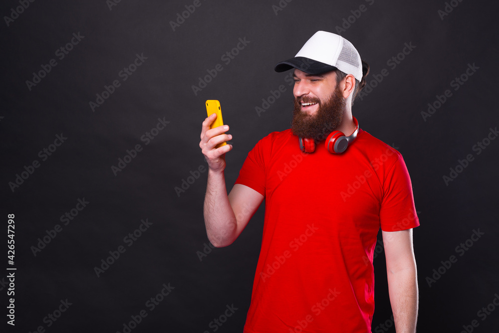 Photo of smiling young man with beard in red t-shirt near black background using smartphone