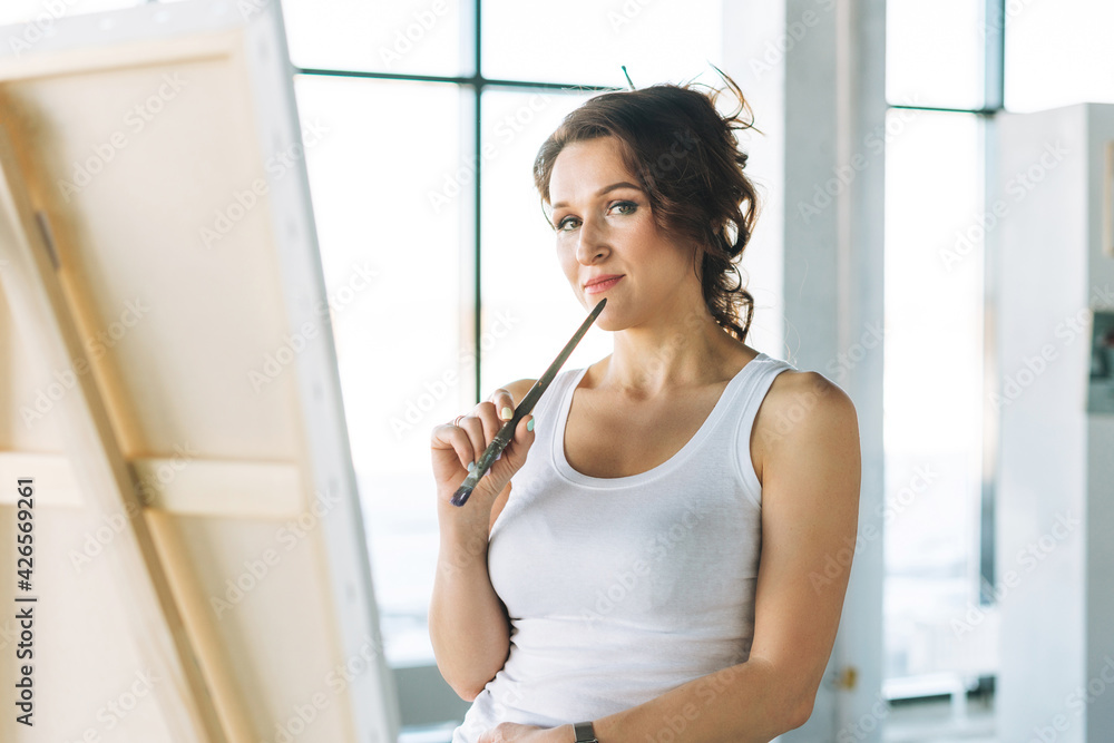 Thinking charming young woman artist in casual clothes with paint brushes in front of easel in modern art gallery Thinking charming young woman artist in casual clothes with paint brushes in front of easel in modern art gallery