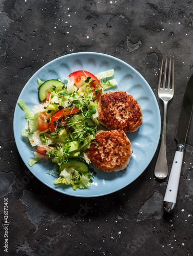 Chicken burgers and fresh iceberg, tomatoes, cucumber salad on a dark background, top view