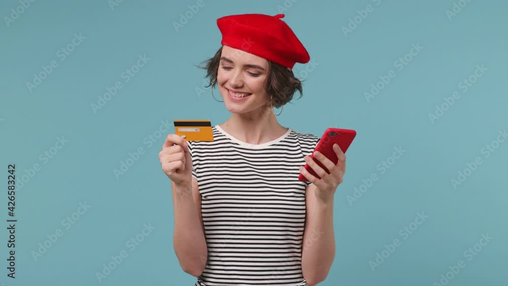 Woman wear red french beret hat striped t-shirt use mobile cell phone hold in hand credit bank card doing online shopping order order delivery to home booking tour isolated on pastel blue background Woman wear red french beret hat striped t-shirt use mobile cell phone hold in hand credit bank card doing online shopping order order delivery to home booking tour isolated on pastel blue background