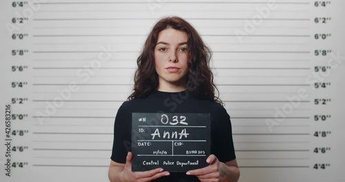 Mugshot of millennial arrested woman with curly hair being photographed in police station. Young female criminal raising head and looking to camera while standing front of metric lineup wall. photo