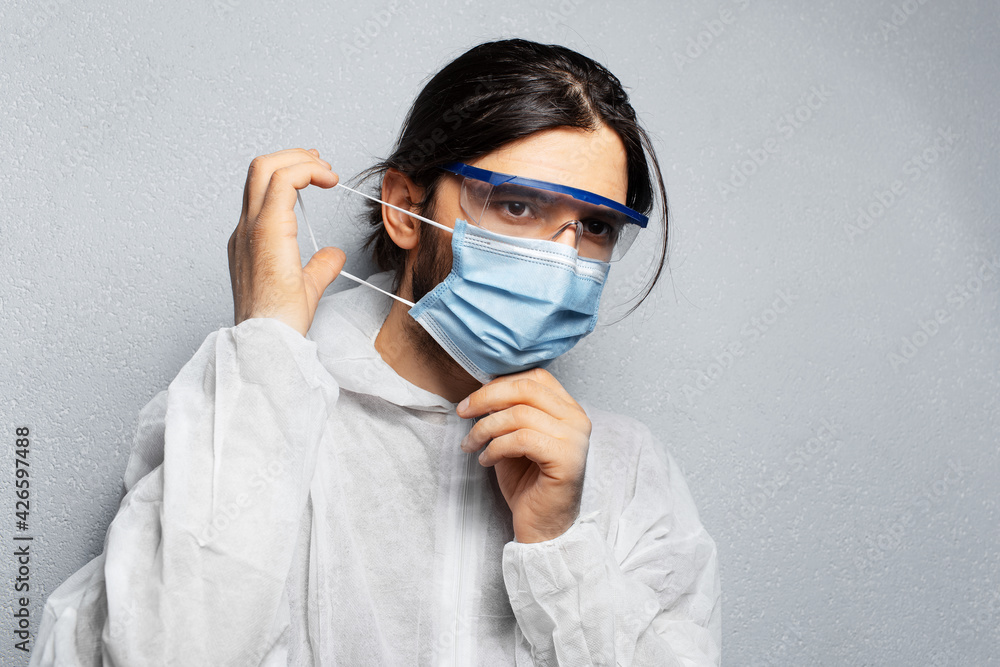 Portrait of young doctor in wearing PPE suit, putting on medical face mask against coronavirus, covid-19. Background of grey wall. Portrait of young doctor in wearing PPE suit, putting on medical face mask against coronavirus, covid-19. Background of grey wall.