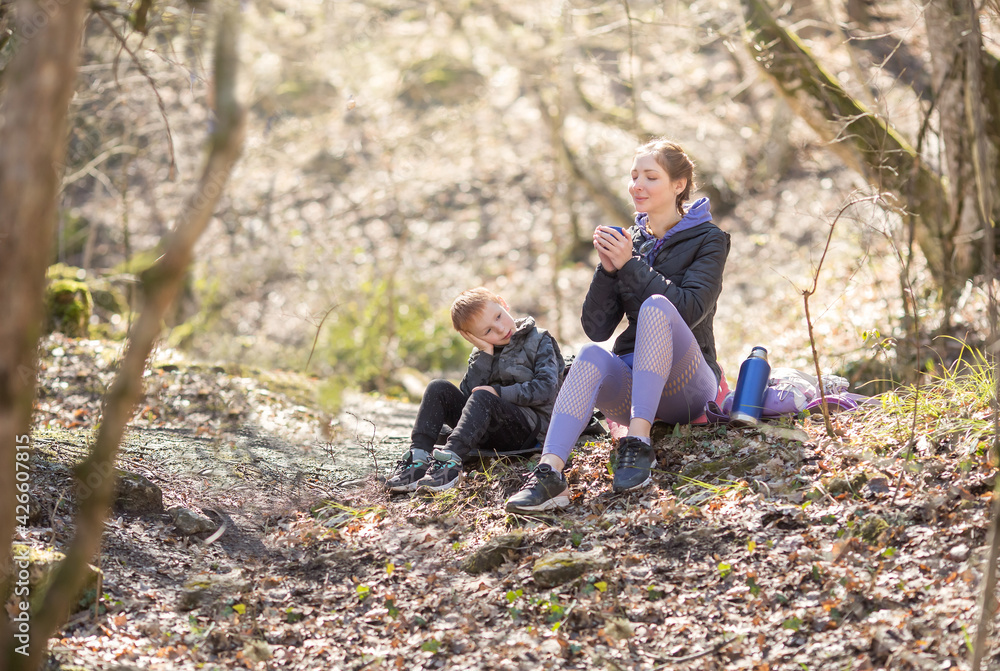 a sports family of tourists a woman and a child sit resting in the forest on a hike, a mother drinks tea a child looks at her