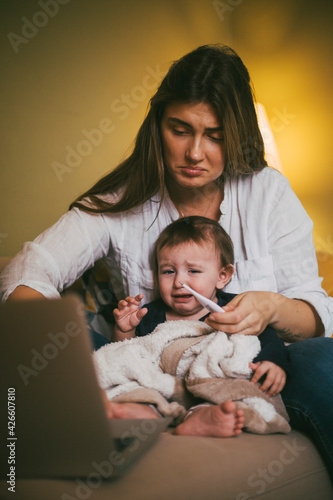 Mother checking temperature of daughter while taking advice online on laptop at home photo