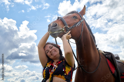 Portrait of beautiful gypsy girl with a horse on a field with green glass in summer day and blue sky and white clouds background. Model in ethnic dress posing with farm animal