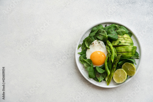 Healthy green breakfast with fried egg. Asparagus, spinach, avocado and egg in plate on white stone background
