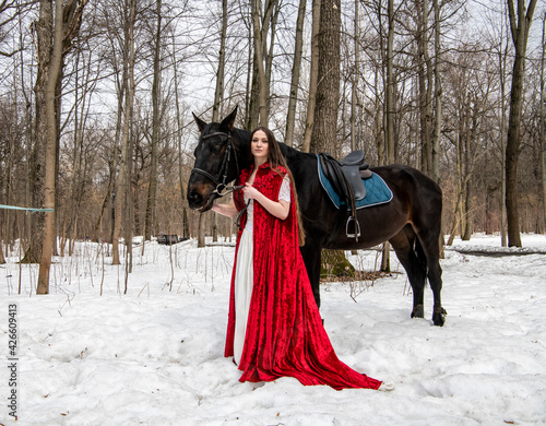 beautiful woman in white dress and red cape with black horse in spring forest 