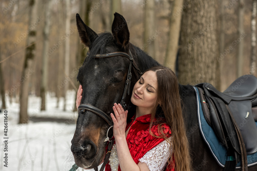 Obraz premium beautiful woman in white dress and red cape with black horse in spring forest 