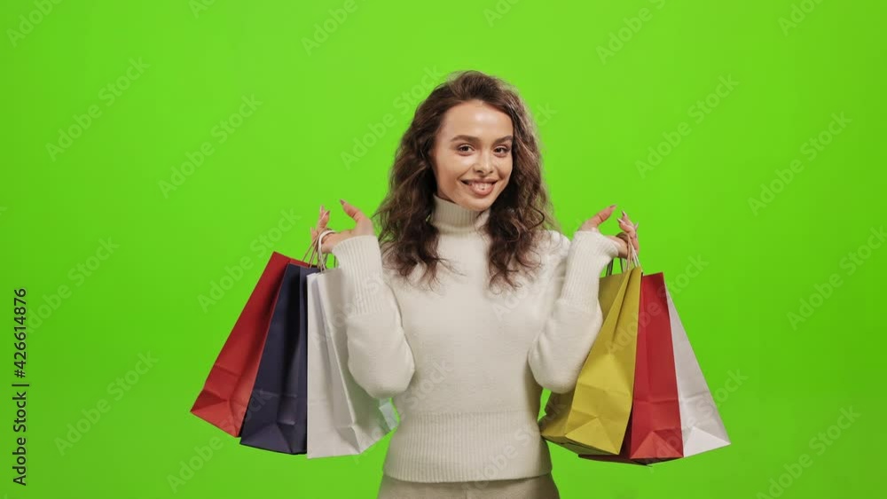 The woman is looking at the camera and smiling. She is picking up the shopping bags. She is standing on a green background. Green screen. 4K The woman is looking at the camera and smiling. She is picking up the shopping bags. She is standing on a green background. Green screen. 4K