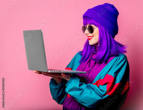 Stylish girl in 80s tracksuit hold laptop on pink background