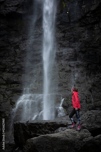Woman stands on rock with magenta jacket, looks at Fossa waterfall on Streymoy Island, Faroe Islands.