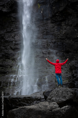 Man stands on rock with red jacket  looks at Fossa waterfall on Streymoy Island  Faroe Islands.