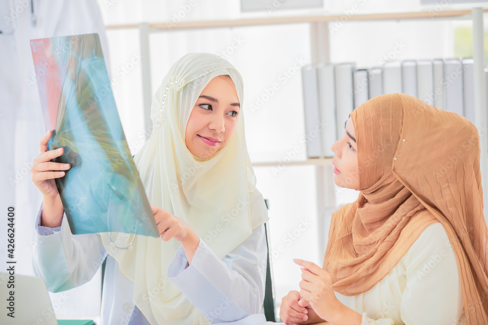 Attractive smiling young adult Muslim doctor wearing cream hijab sitting and pointing the chest x-ray film to show and explain the abnormalities of lungs to patient Attractive smiling young adult Muslim doctor wearing cream hijab sitting and pointing the chest x-ray film to show and explain the abnormalities of lungs to patient