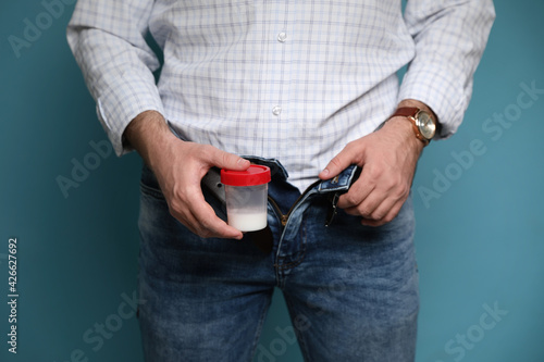 Donor with unzipped pants holding container of sperm on turquoise background, closeup